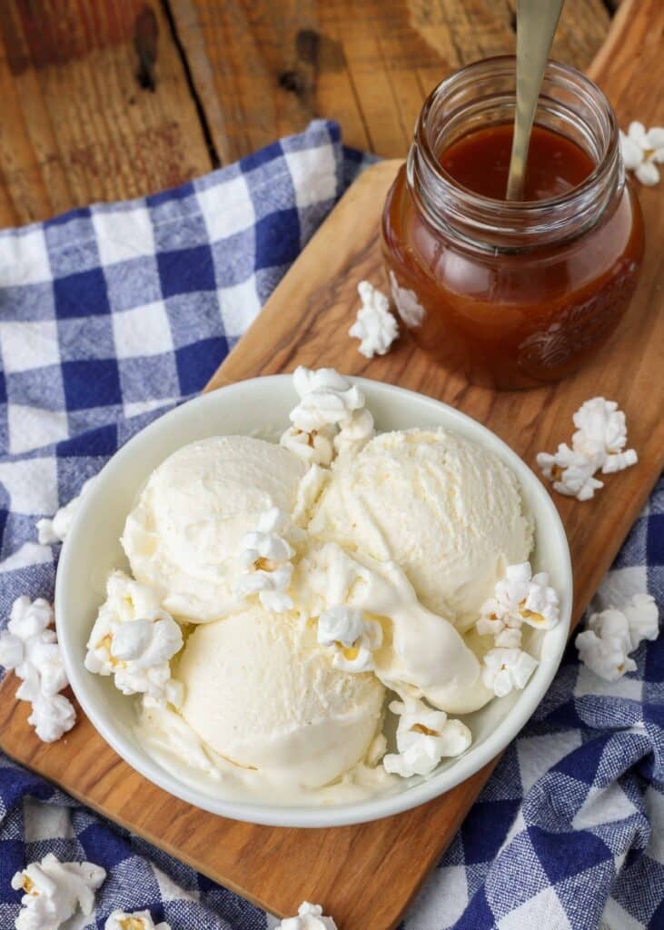 a top down photo of a serving board with a bowl of ice cream and a jar of caramel sauce on it, with a blue and white checkered napkin in the background.
