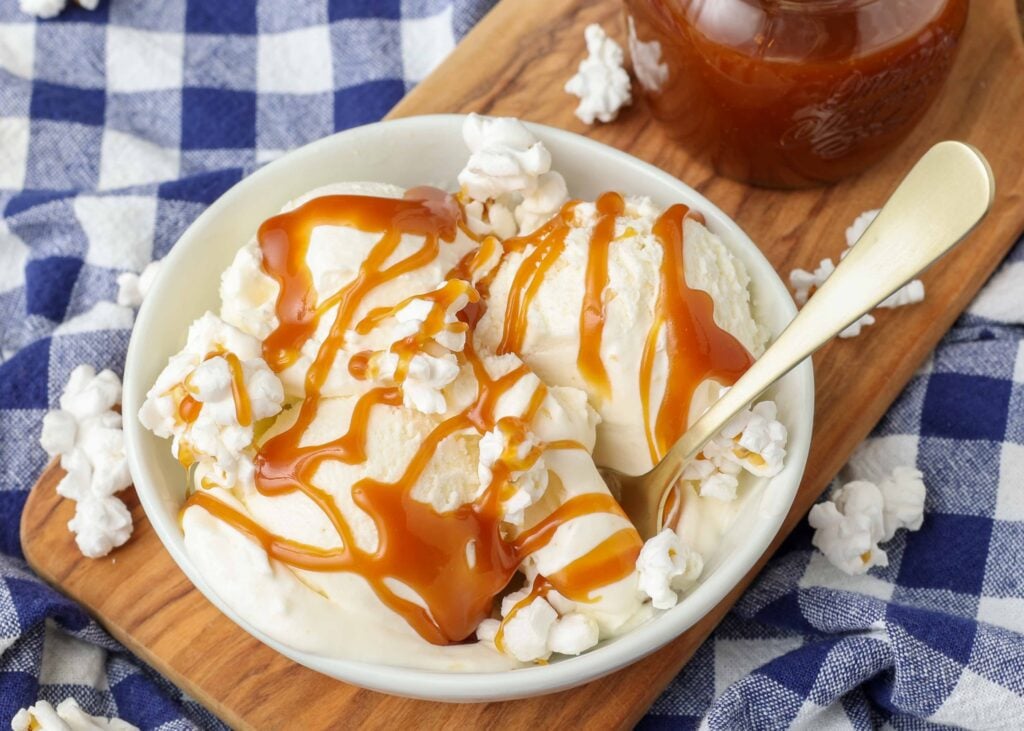 a horizontally aligned photo of a bowl of popcorn ice cream that has been drizzled with caramel sauce, and a metal spoon handle sticking out of it