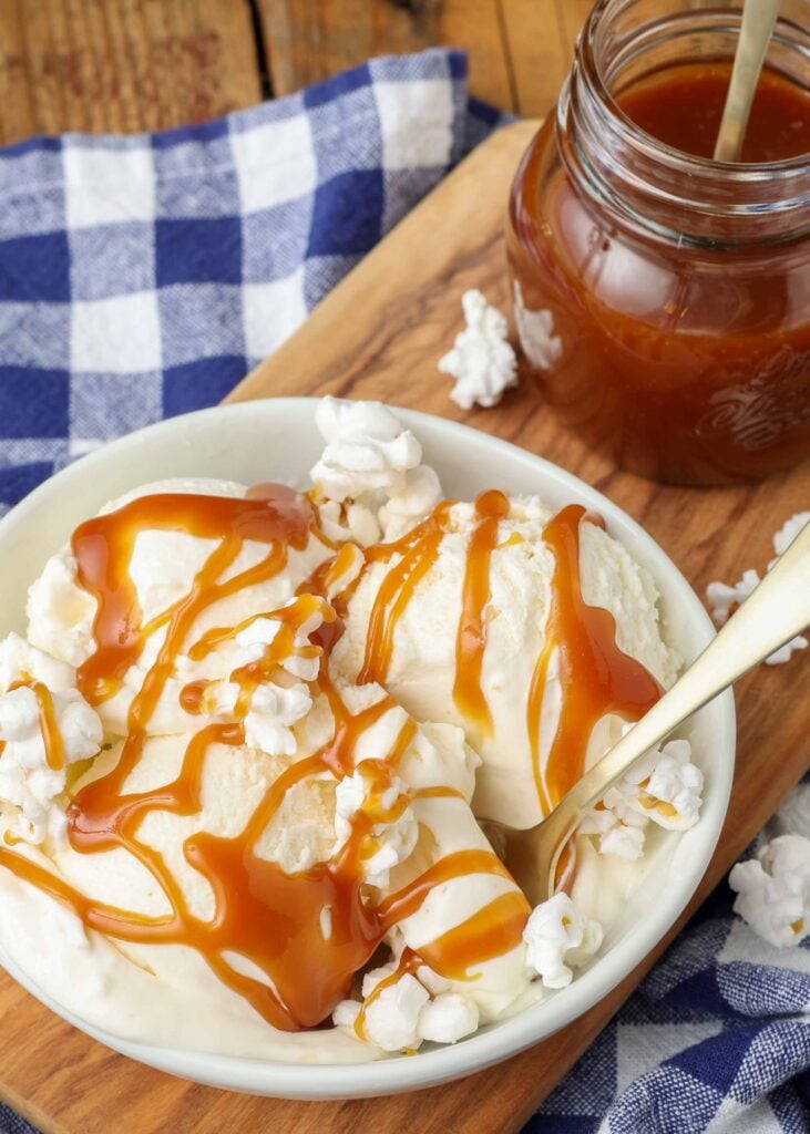 a vertically aligned photo of a bowl of popcorn ice cream that has been drizzled with caramel sauce, and a metal spoon handle sticking out of it