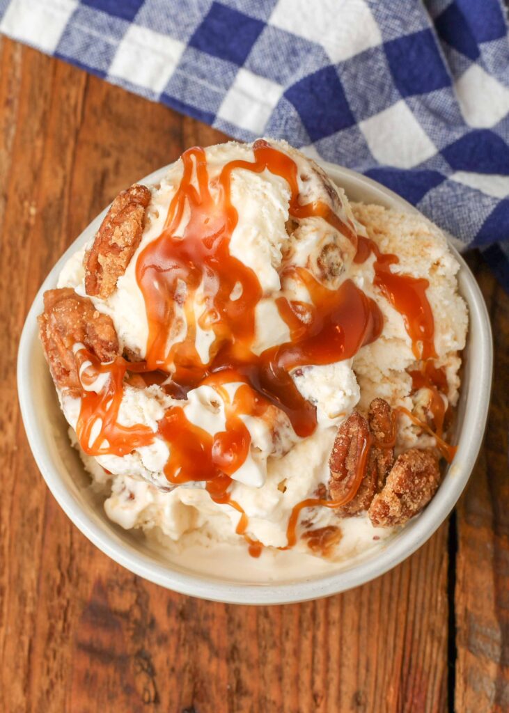 Overhead shot of pralines and cream ice cream topped with pecans and caramel sauce, served in a white bowl with a checkered blue and white towel