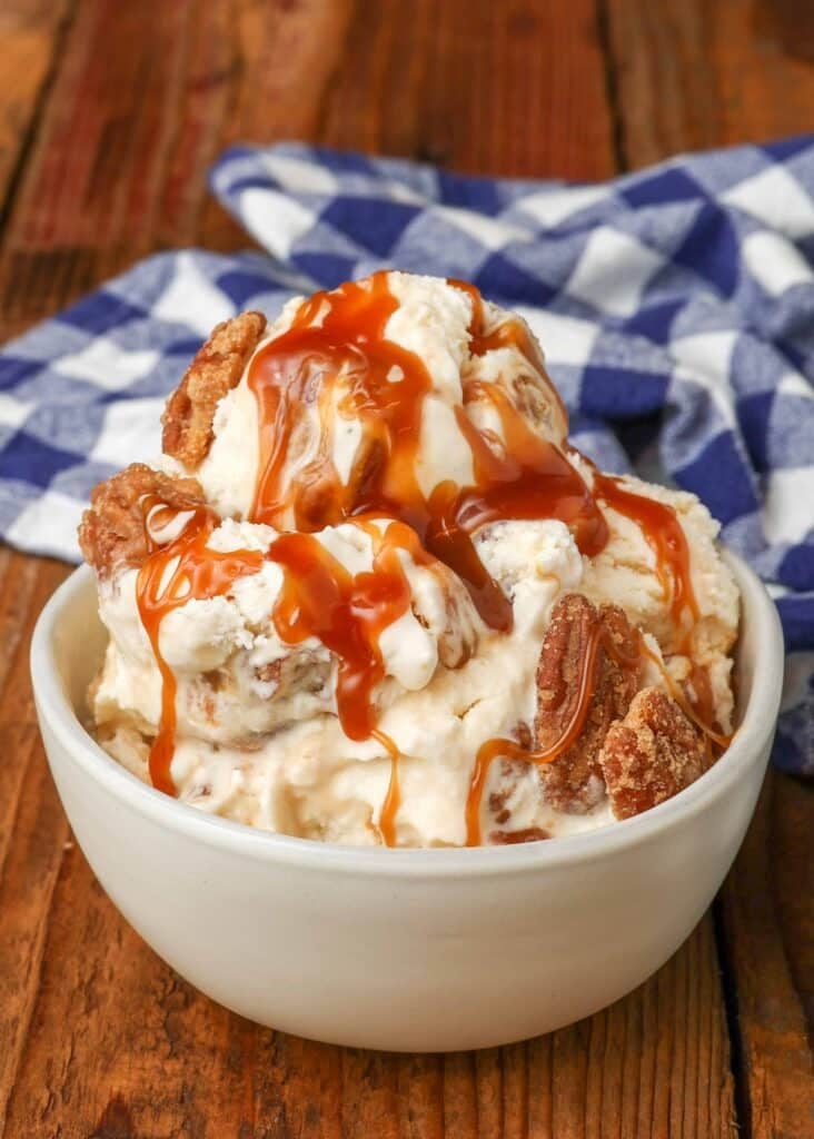 Vertical shot of pralines and cream ice cream topped with pecans and caramel sauce, served in a small white bowl with a checkered blue and white towel