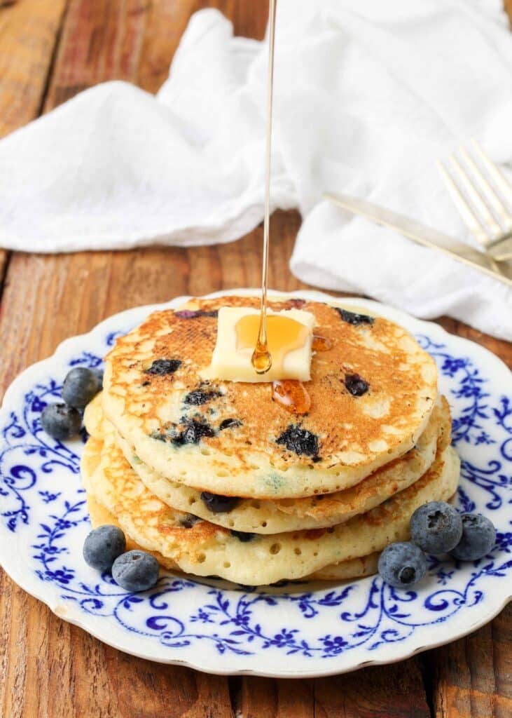 pouring maple syrup over a pat of butter atop a pile of buttermilk blueberry pancakes