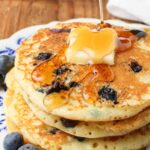 A tropical up shot of drizzling maple syrup over a pat of butter atop a pile of buttermilk blueberry pancakes