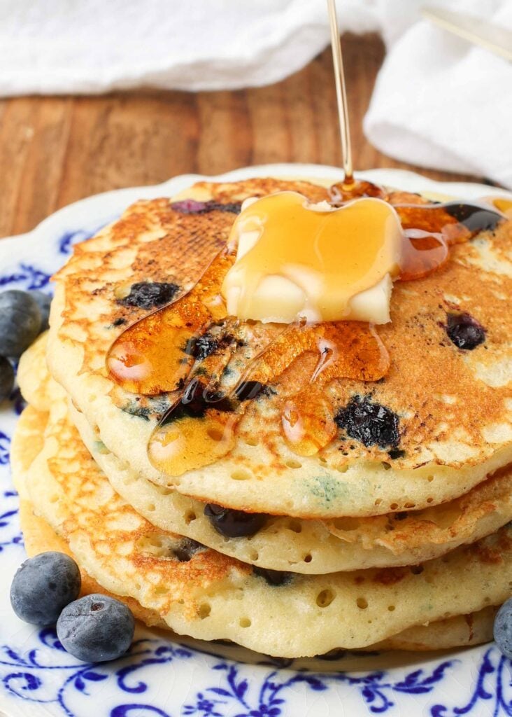 A close up shot of drizzling maple syrup over a pat of butter atop a pile of buttermilk blueberry pancakes