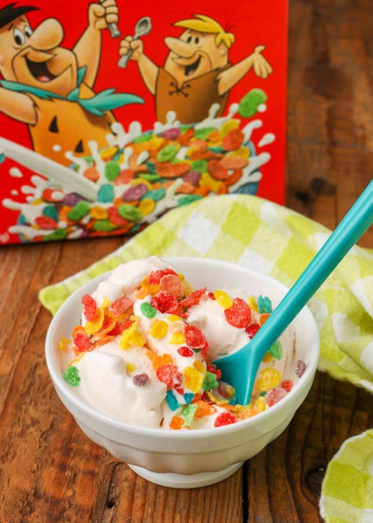 Overhead shot of Fruity Pebbles Ice Cream topped with Fruity Pebbles cereal, served in a small white bowl with a small blue plastic spoon
