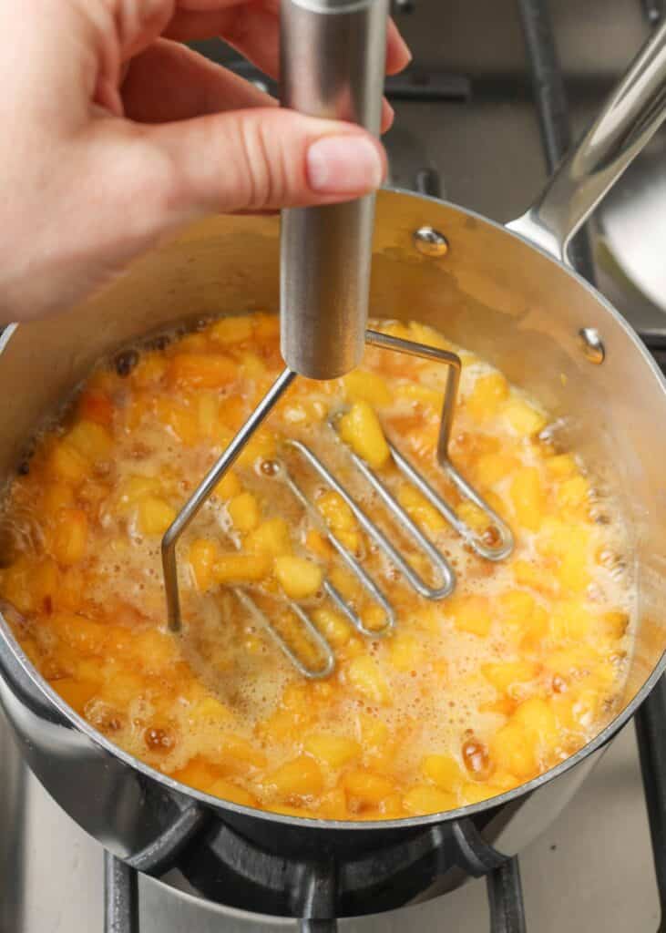 Overhead shot of peaches cooking in stainless steel pan, with a metal potato masher