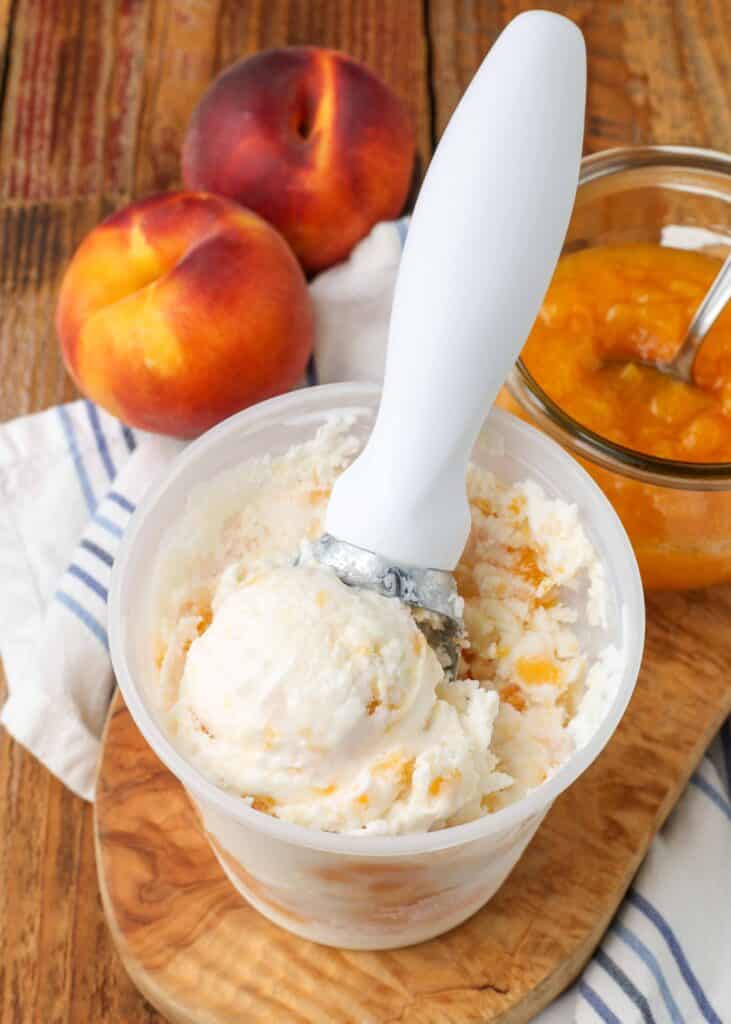 Overhead shot of peaches and cream ice cream, stored in a white plastic tub with a white ice cream scoop