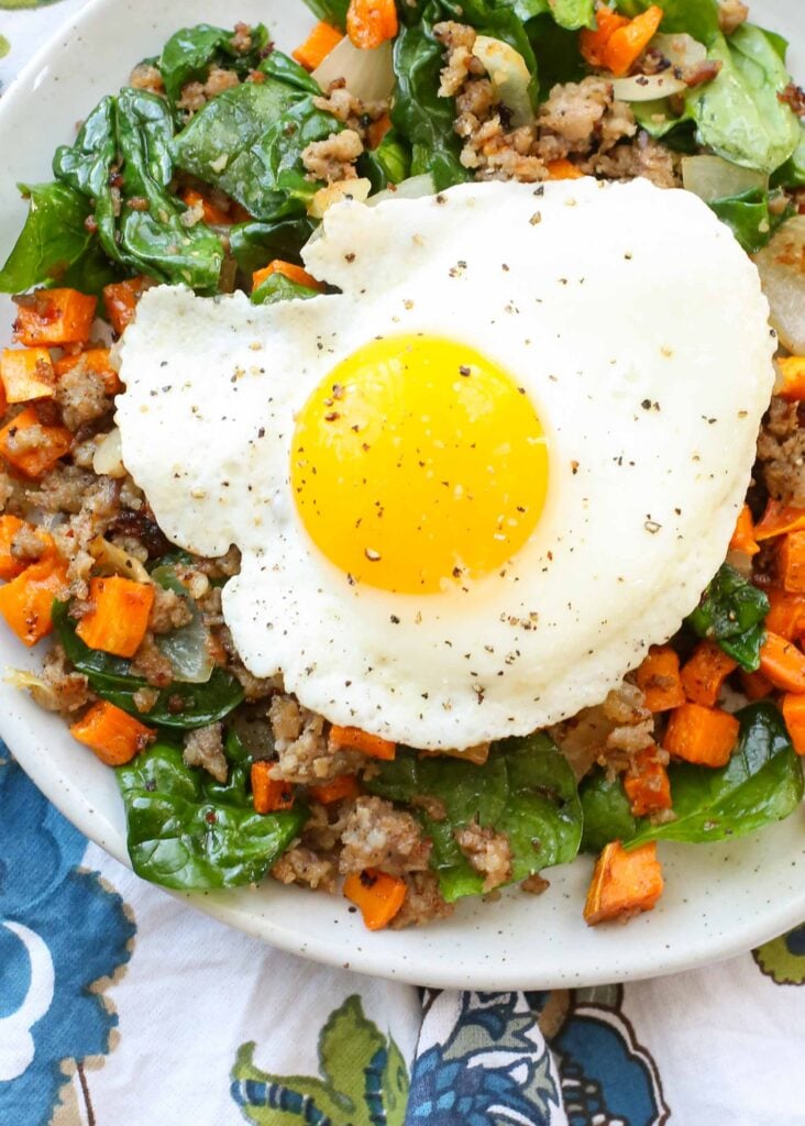 Overhead horizontal shot of sausage sweet potato hash topped with a sunny side up egg, served in a white bowl with a blue and white floral pattern hand towel