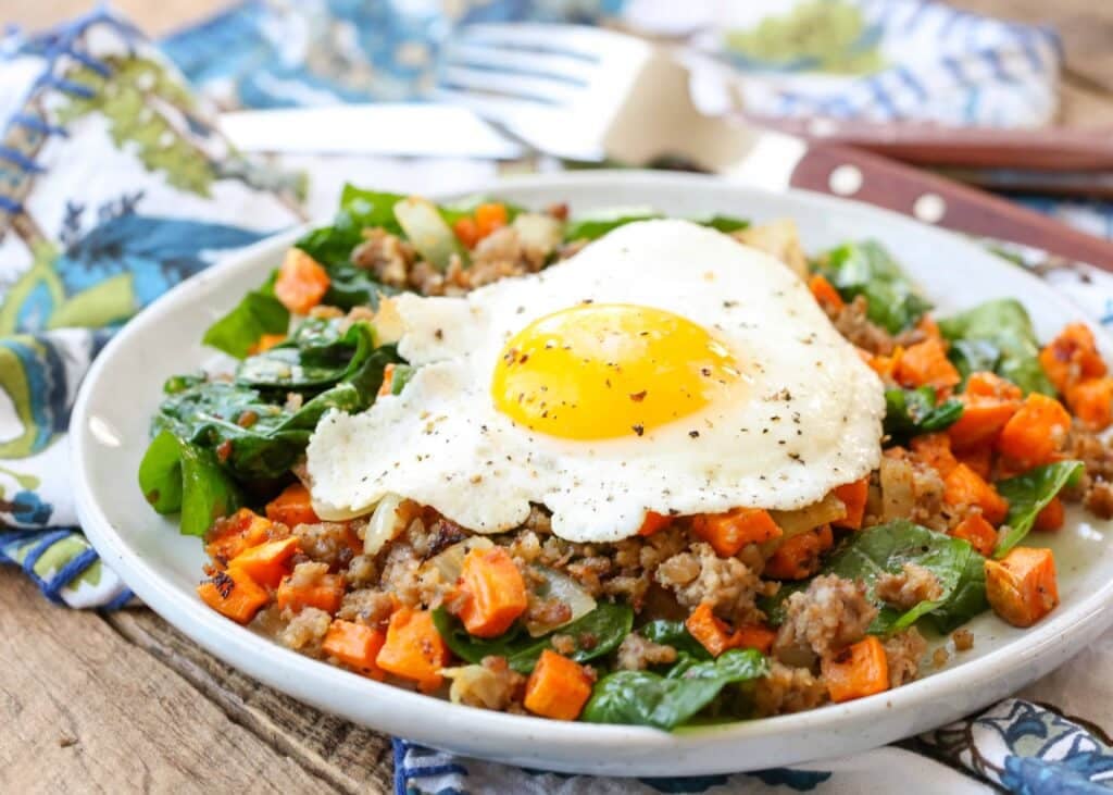 Horizontal shot of sausage sweet potato hash with a sunny side up egg, served in a white bowl with a blue and white floral pattern towel