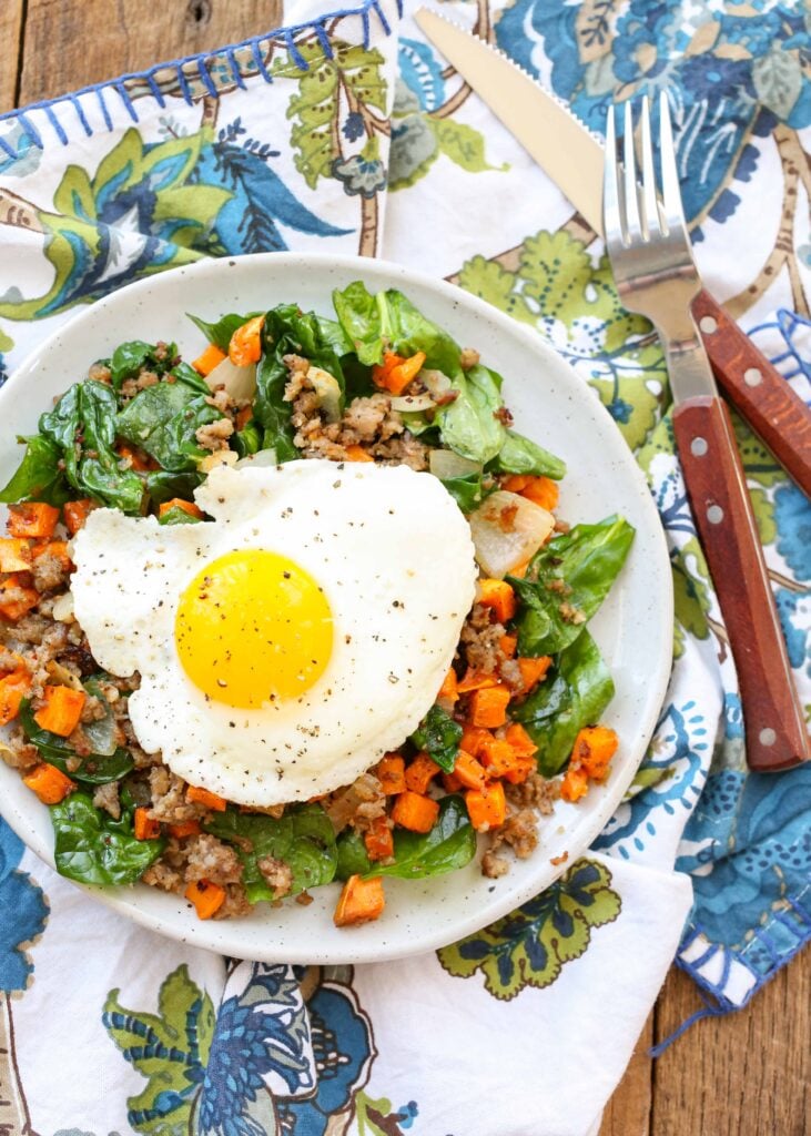 Overhead shot of sausage sweet potato hash with a sunny side up egg, served in a white bowl with a blue and white floral pattern towel