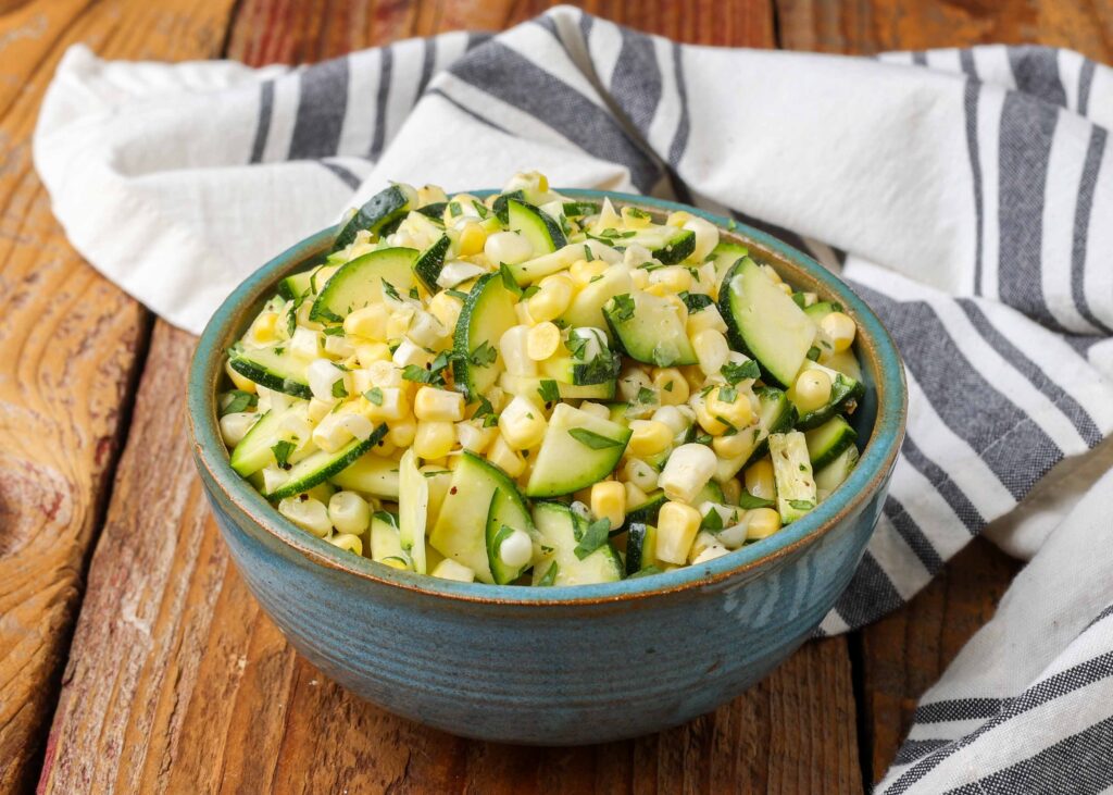 Horizontal shot of zucchini corn salad, served in a small blue ceramic bowl with a striped white and gray towel