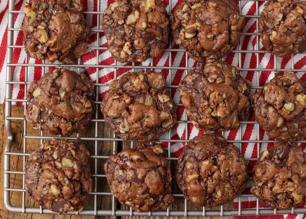 Overhead horizontal close-up shot of chocolate walnut cookies on a silver cooling rack; underneath, a striped red and white hand towel
