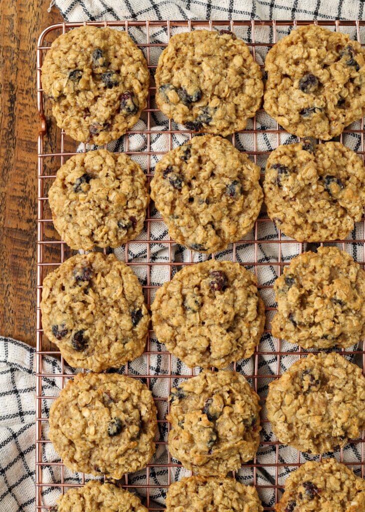 Overhead vertical shot of cranberry orange oatmeal cookies on a copper cooling rack; underneath, a checkered white and black hand towel