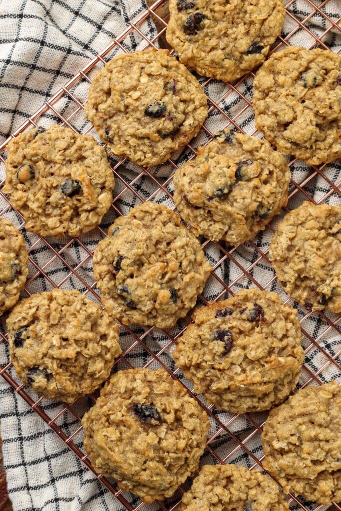 Overhead zoomed-out vertical shot of cranberry orange oatmeal cookies on a copper cooling rack; underneath, a checkered white and black hand towel