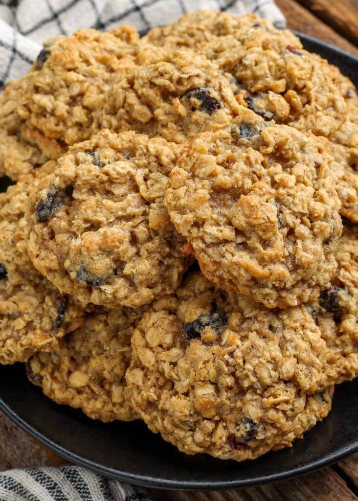 Overhead close-up vertical shot of cranberry orange oatmeal cookies, piled in a black bowl with a checkered white and black hand towel