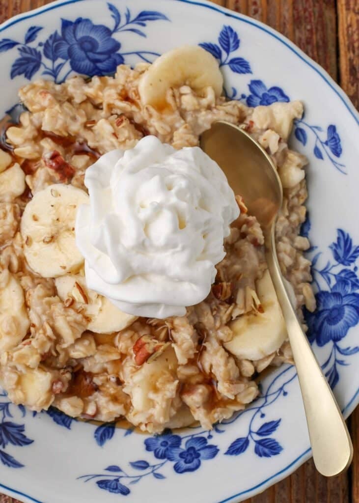 Overhead close-up shot of maple banana nut oatmeal topped with whipped cream, served with a silver spoon in a white bowl with blue floral print