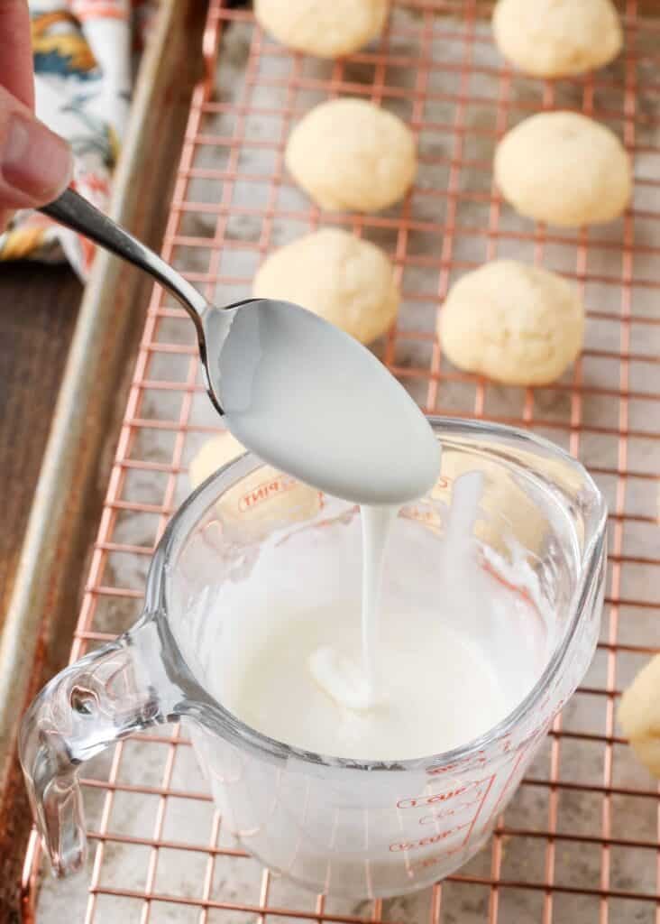 a spoon drizzling glaze over a measuring cup next to small almond butter cookies on a cooling rack over a sheet pan