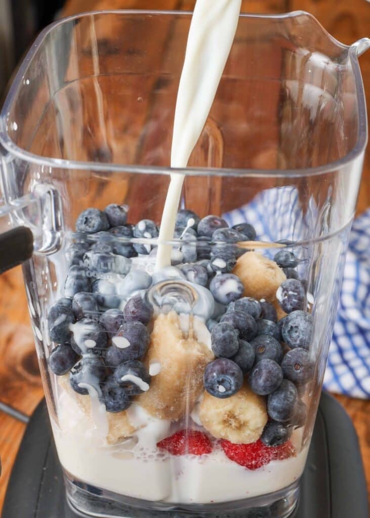 Pouring milk into blender pitcher filled with raspberries, blueberries, and bananas
