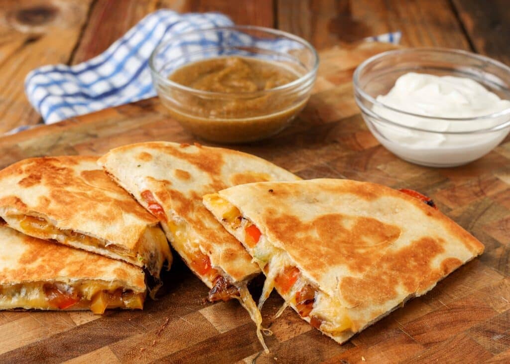 Stacked wedges of fajita quesadilla on a wooden board with a blue and white checkered tea towel in the background.