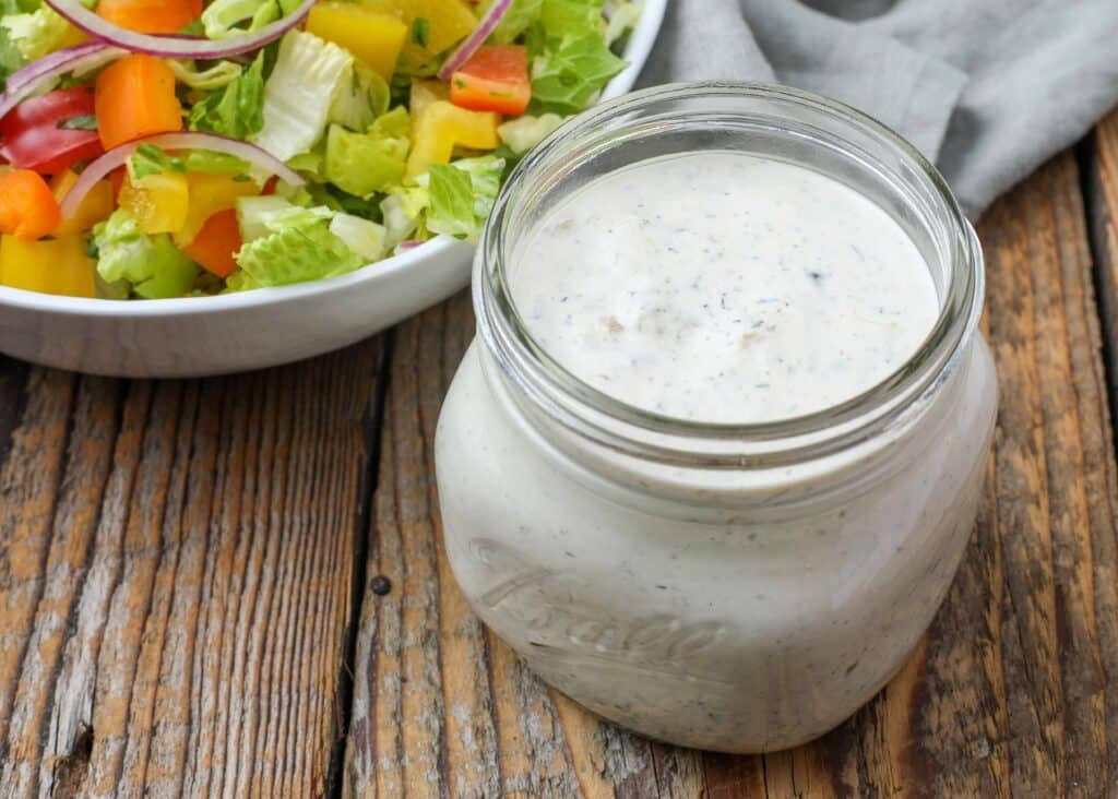 close up overhead photo of creamy salad dressing in jar next to salad