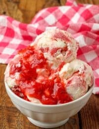 Strawberry lemonade ice cream scooped into a bowl with a drizzle of strawberry sauce and red and white tea towel on the side.