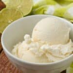 Vertical close-up shot of fluffy off-white ice cream in a small white bowl; in the background, halved limes rest on a light green towel
