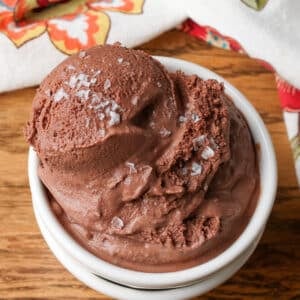 Chocolate sea salt ice cream has been scooped into a white serving dish over a wooden tabletop in this vertically aligned image.