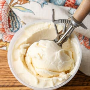 A picture of a freezer safe container full of luscious ice cream on a wooden tabletop. A wooden handled ice cream scoop has a ball of ice cream in the foreground.