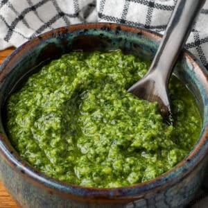 A close up shot of pesto in a bowl with a black spoon. A black and white checkered tea towel is visible in the background.