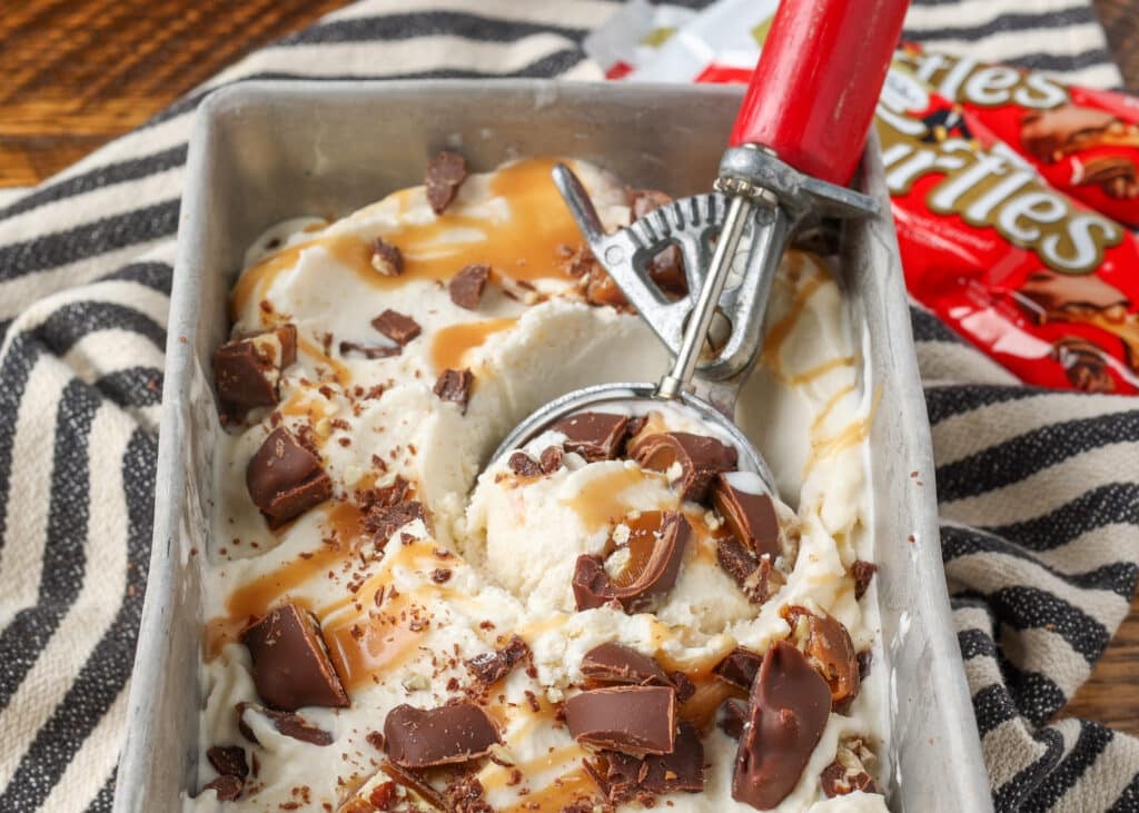 A horizontally aligned image of an ice cream scoop with a red handle in a freezer safe tin full of this luscious dessert.