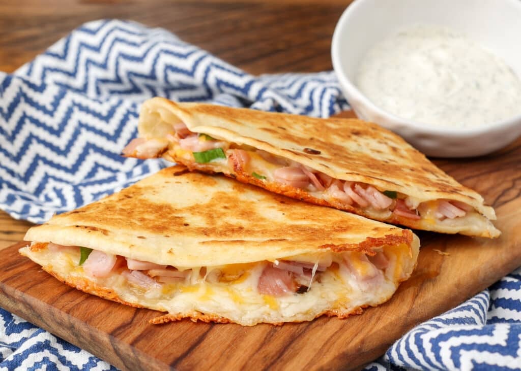 A ham and cheese quesadilla has been sliced and is ready to eat, served on a wooden board with a side of dip!