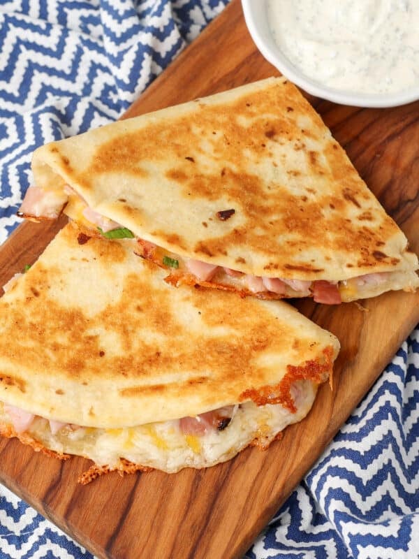 A vertically aligned image of a ham and cheese quesadilla that has been sliced and is ready to eat, served on a wooden board with a side of dip!