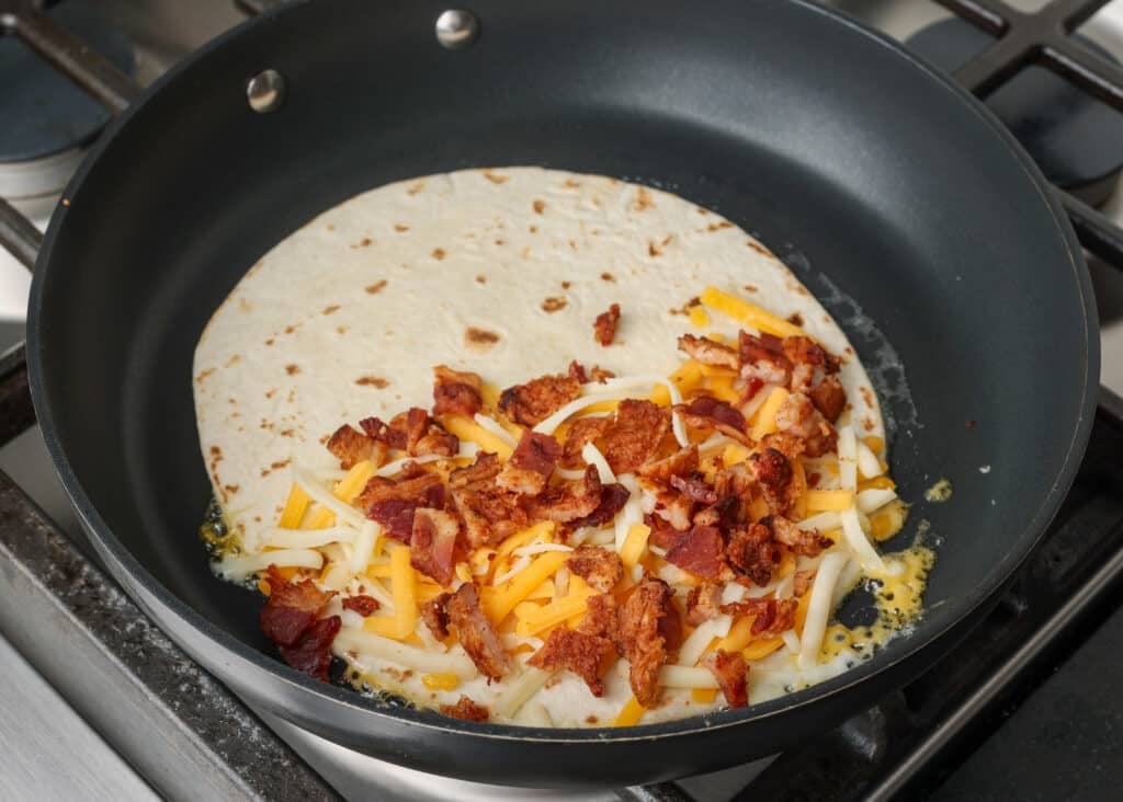 the first layer of cheese and bacon have been added to half the quesadilla in a non-stick pan