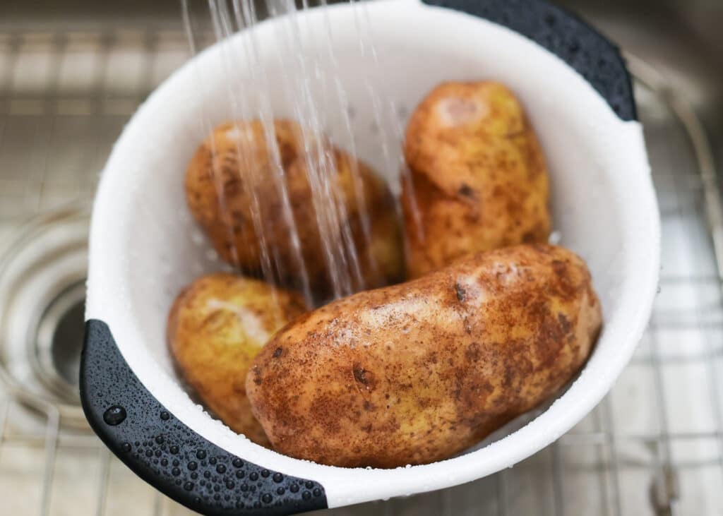 Cleaning the potatoes in a white colander under the faucet with cold water.