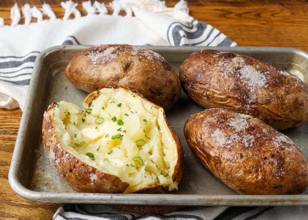 Perfectly baked potatoes from the oven are on a rimmed baking sheet, and one has been split open and covered with fresh herbs and melting butter. 