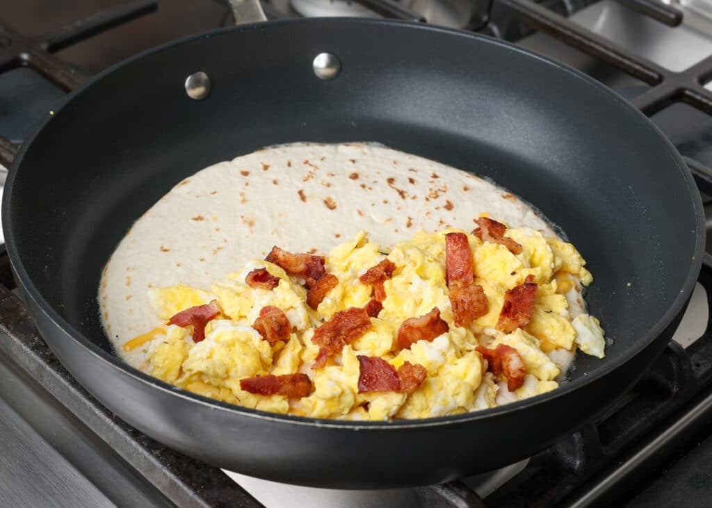 One half of the tortilla has been piled high with cheese, eggs, and bacon in the pan.