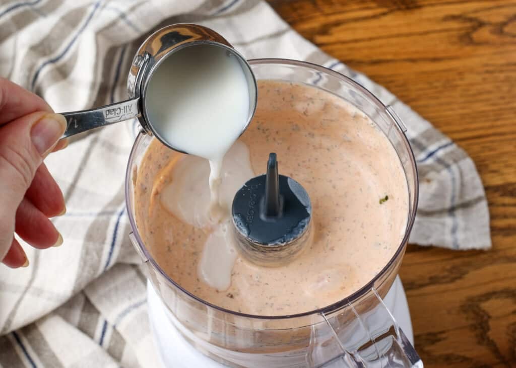 A measuring cup pours white liquid into the food processor to make the creamy dressing.