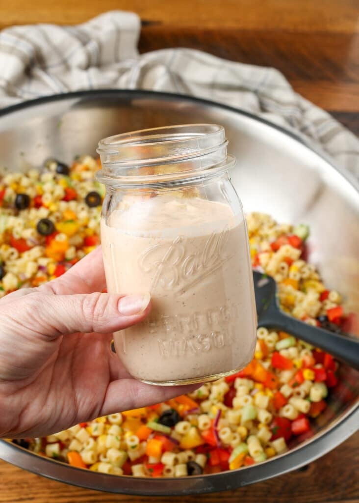 A vertically aligned image of a hand holding the dressing over the salad ingredients in a metal mixing bowl.