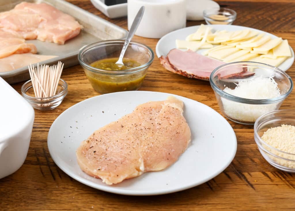 A raw chicken cutlet has been laid out on a plate beside the other ingredients for cordon bleu.