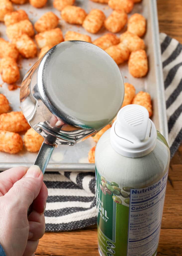 A vertically aligned action shot showing the olive oil spray coating the bottom of the flat-bottomed measuring cup.