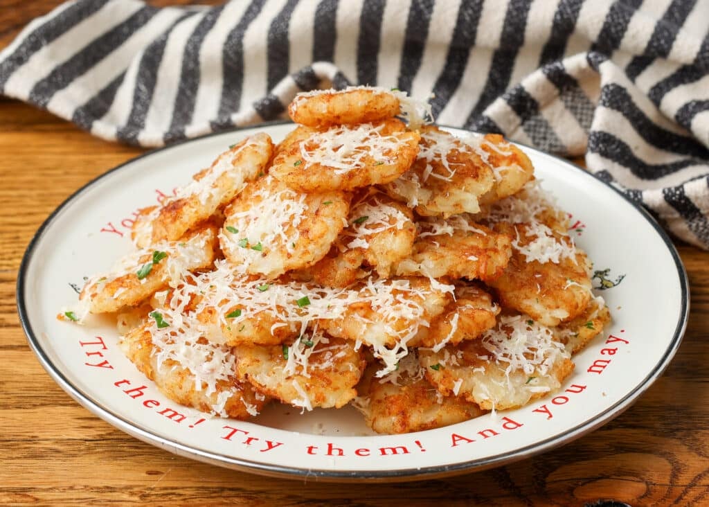 A horizontally aligned image of a white plate on a wooden tabletop with a black and white tea towel visible in the background at the top. Crispy tater tots are piled high on the plate, sprinkled with fresh chopped parsley and shredded parmesan.