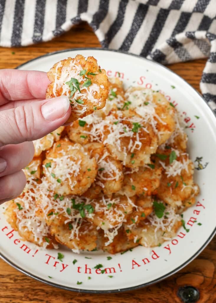 A vertically aligned image with a hand in the foreground, holding a smashed tater tot covered with shredded parmesan and chopped parsley.