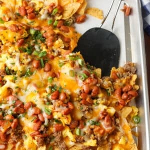 A vertically aligned photo of a baking sheet covered with crisp tortilla chips, meat, beans, cheese, and other toppings.
