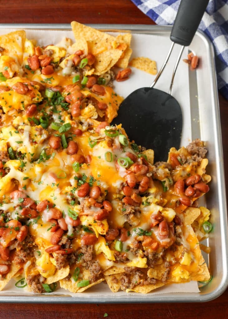A vertically aligned photo of a baking sheet covered with crisp tortilla chips, meat, beans, cheese, and other toppings.