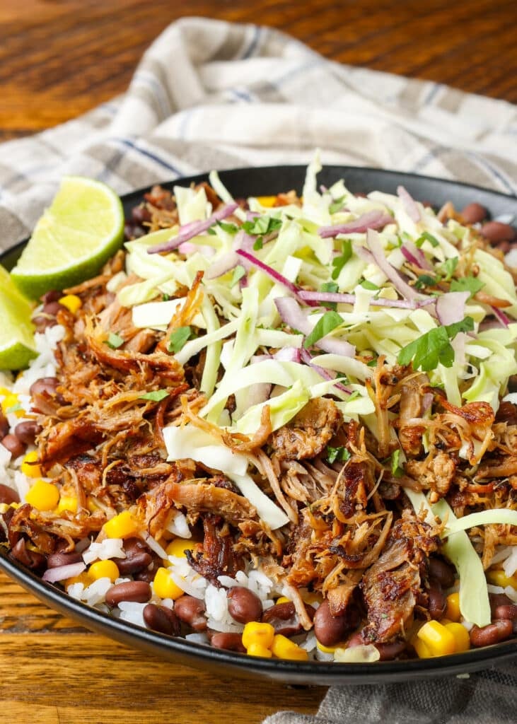 A vertically aligned image showing a bowl loaded with fresh toppings and warm ingredients on a wooden tabletop with a grey and white tea towel visible in the background.