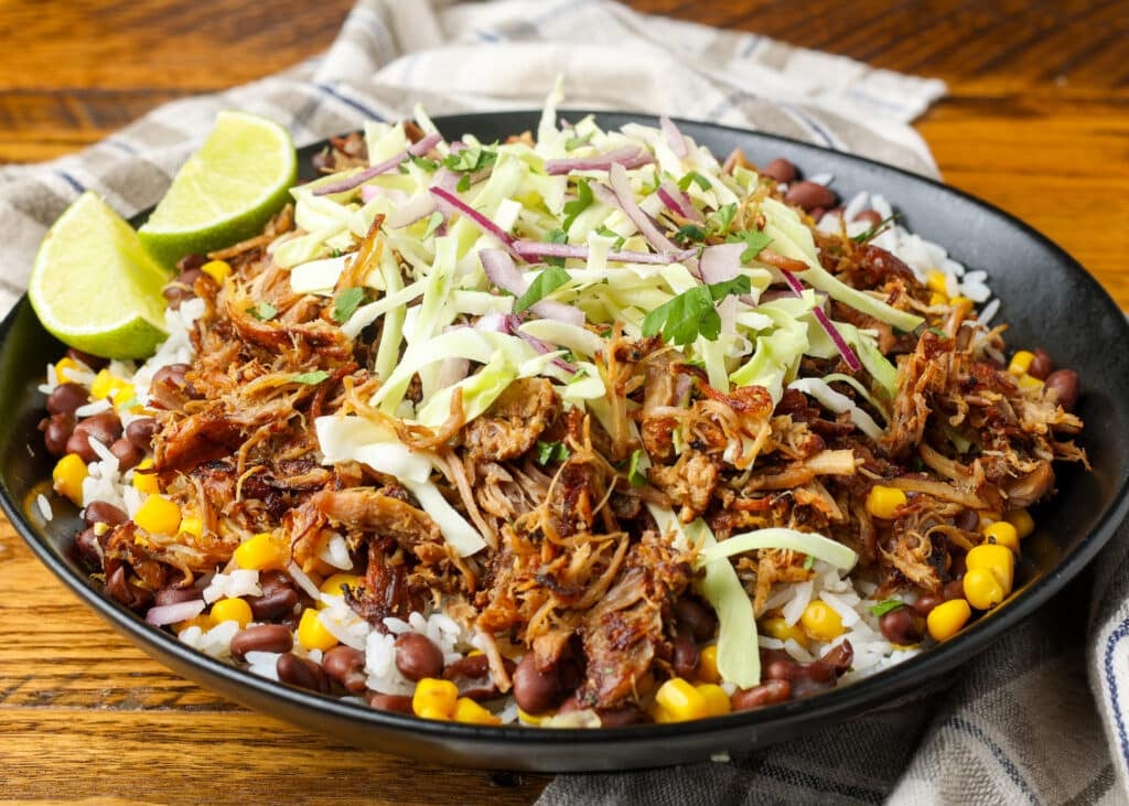 A horizontally aligned image of a pork carnitas bowl piled high with cabbage, rice, beans, corn, limes, and avocadoes, ready to eat!