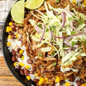 A vertically aligned top-down image of a carnitas bowl loaded high with pork, rice, beans, corn, and cabbage with lime wedges on the side.