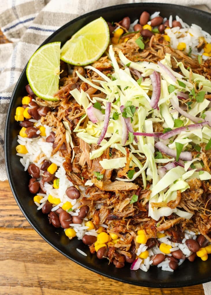 A vertically aligned top-down image of a carnitas bowl loaded high with pork, rice, beans, corn, and cabbage with lime wedges on the side.