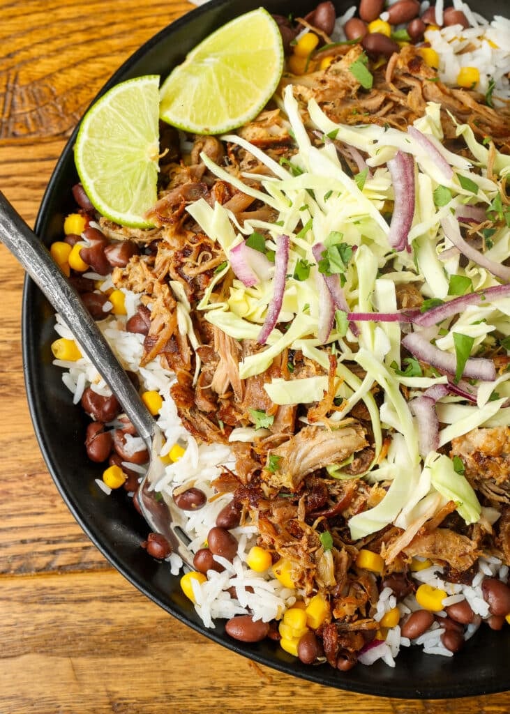 A vertically aligned, top down close up image of a bowl ready to eat, with a black metal fork sticking out. Also visible are the lime wedgeson top of the bowl at the edge, and the wooden tabletop in the background.
