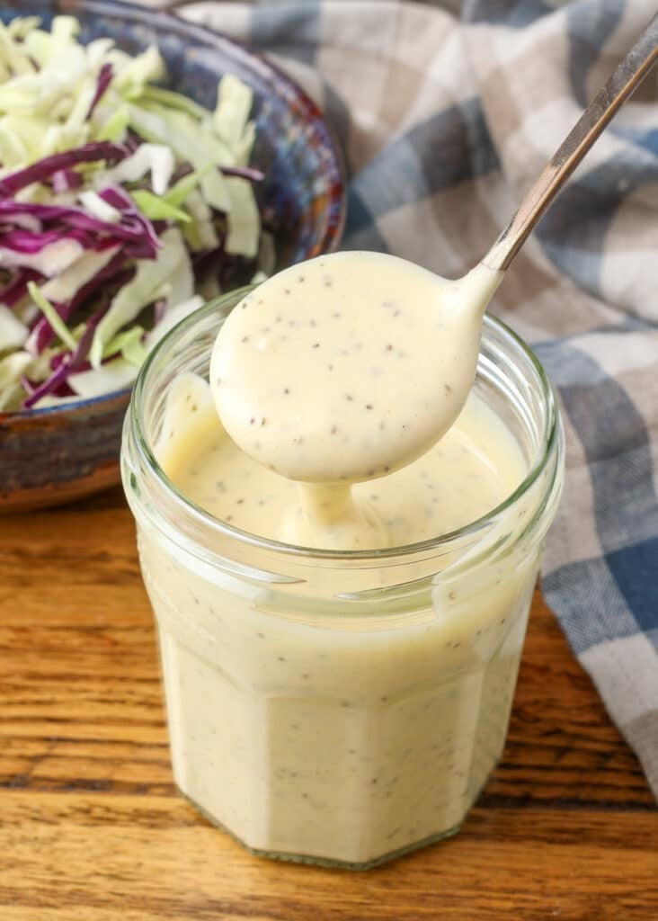 A vertically aligned close up photo showing the dressing in a glass jar over a wooden tabletop with the shredded cabbage in a bowl visible in the upper left background.